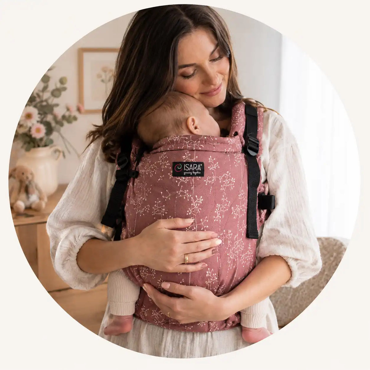 Mãe a transportar o bebé no marsúpio ergonómico ISARA The One rosa com padrão floral, num ambiente acolhedor.