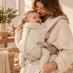 Mãe com bebé na mochila porta-bebé Voksi Huddle bege, num ambiente lifestyle acolhedor com tons suaves e decoração natalícia discreta.